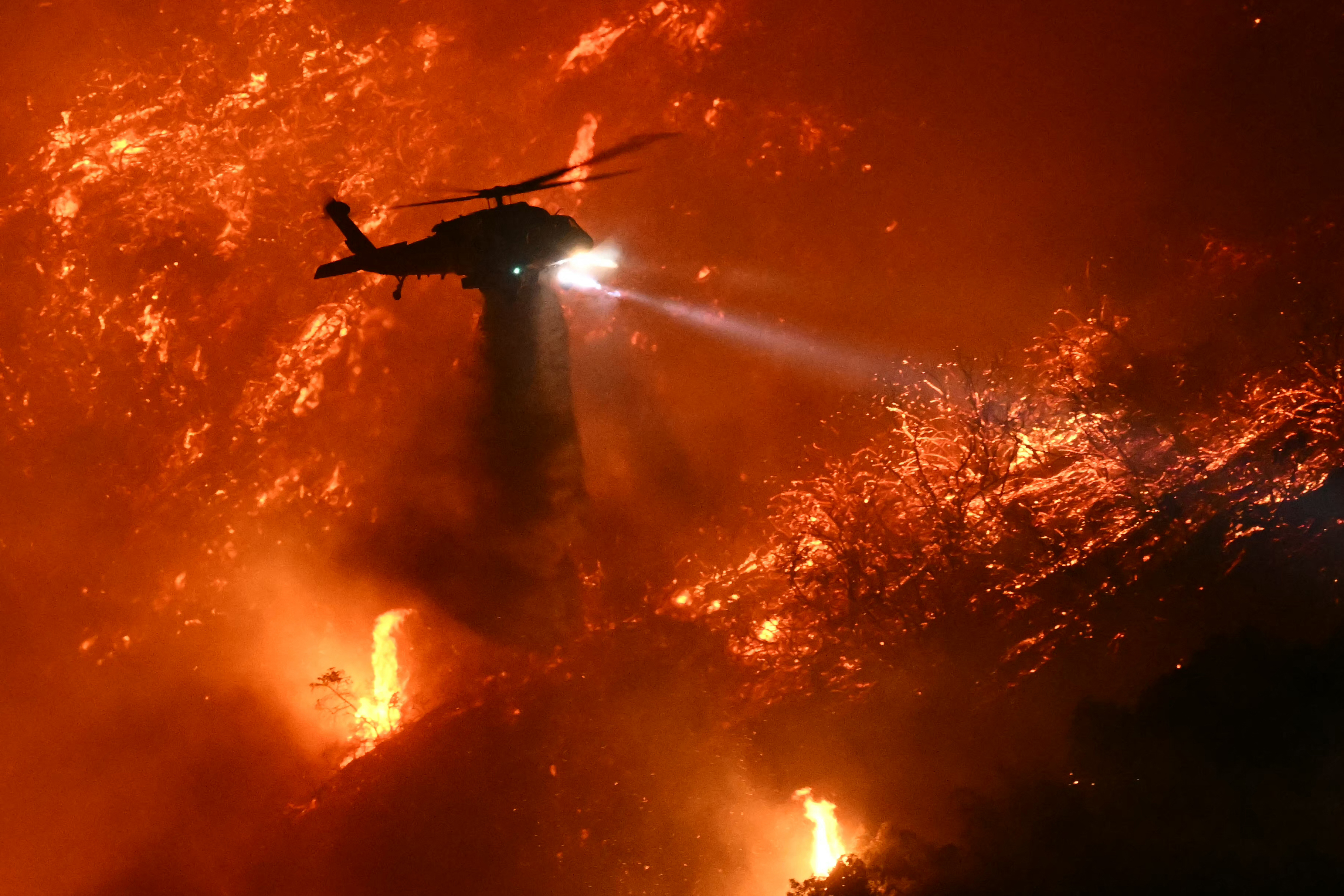A firefighting helicopter drops water as the Palisades fire grows near the Mandeville Canyon neighbourhood and Encino, California. Photo / AFP