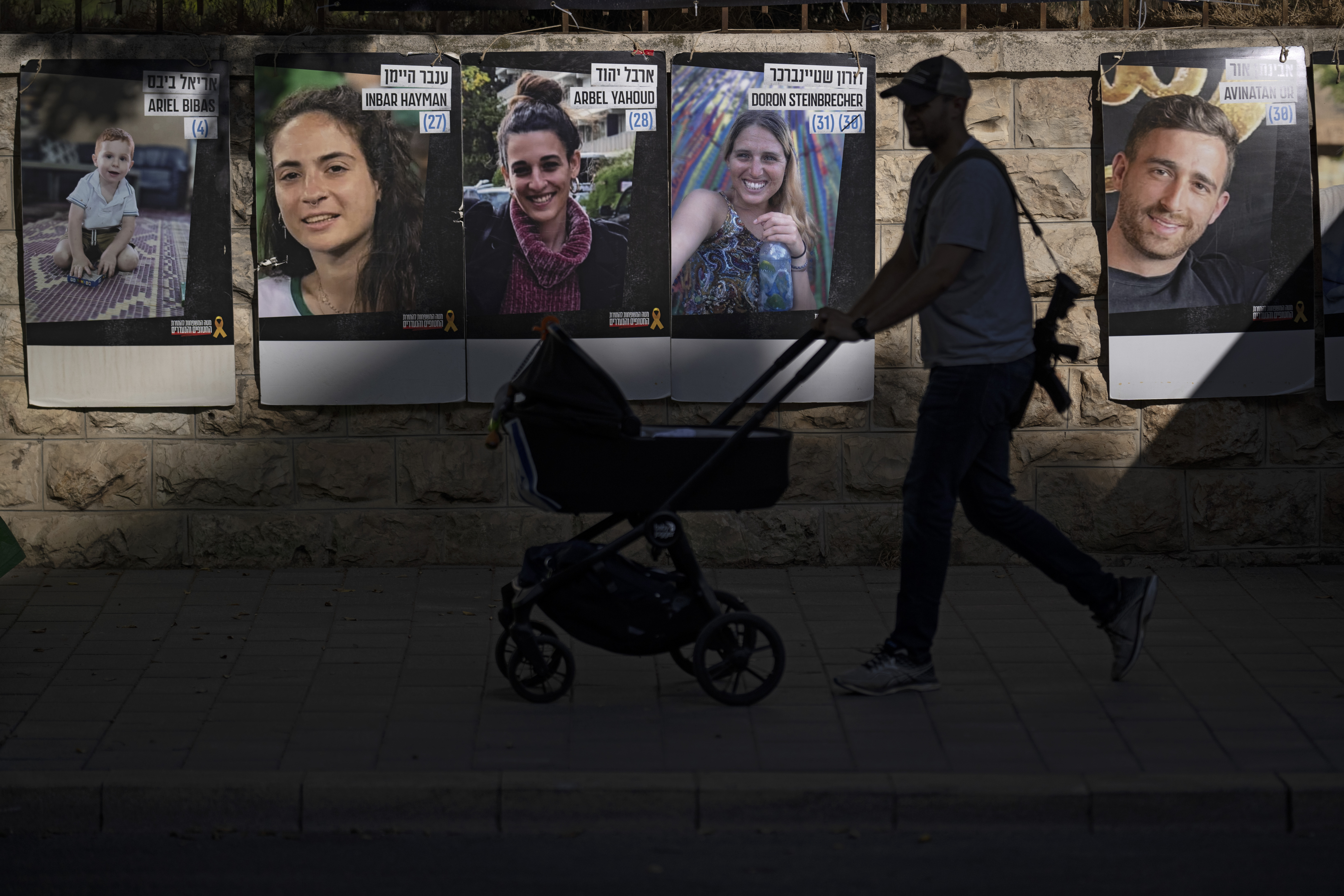 Carrying an weapon, a man in Jerusalem pushes a baby trolley past a wall with photographs of hostages. Photo / Ap