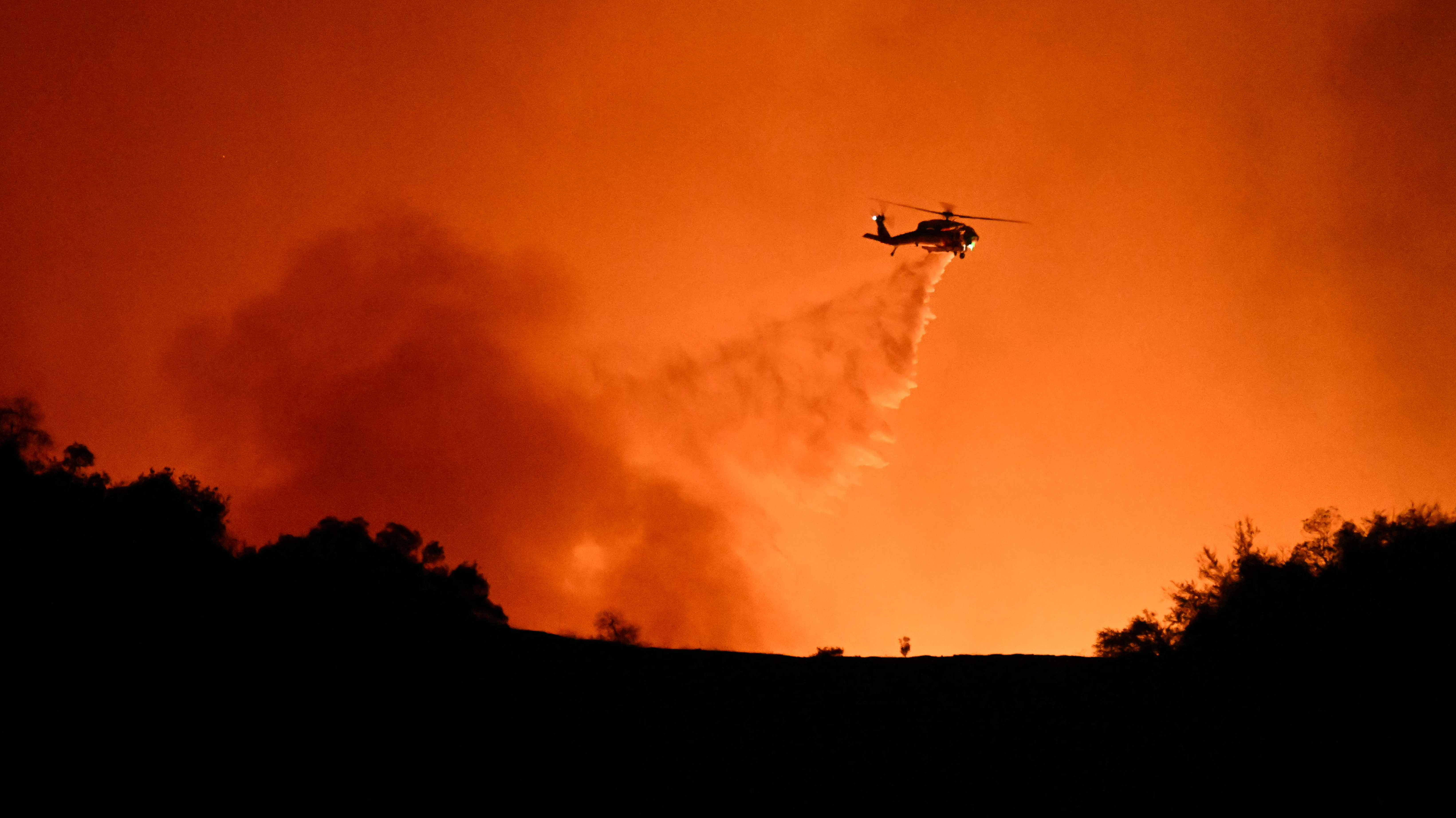 A helicopter drops water on smoke and flames from the Palisades fire burning behind Mulholland Drive towards the Encino neighbourhood in Los Angeles. Photo / AFP