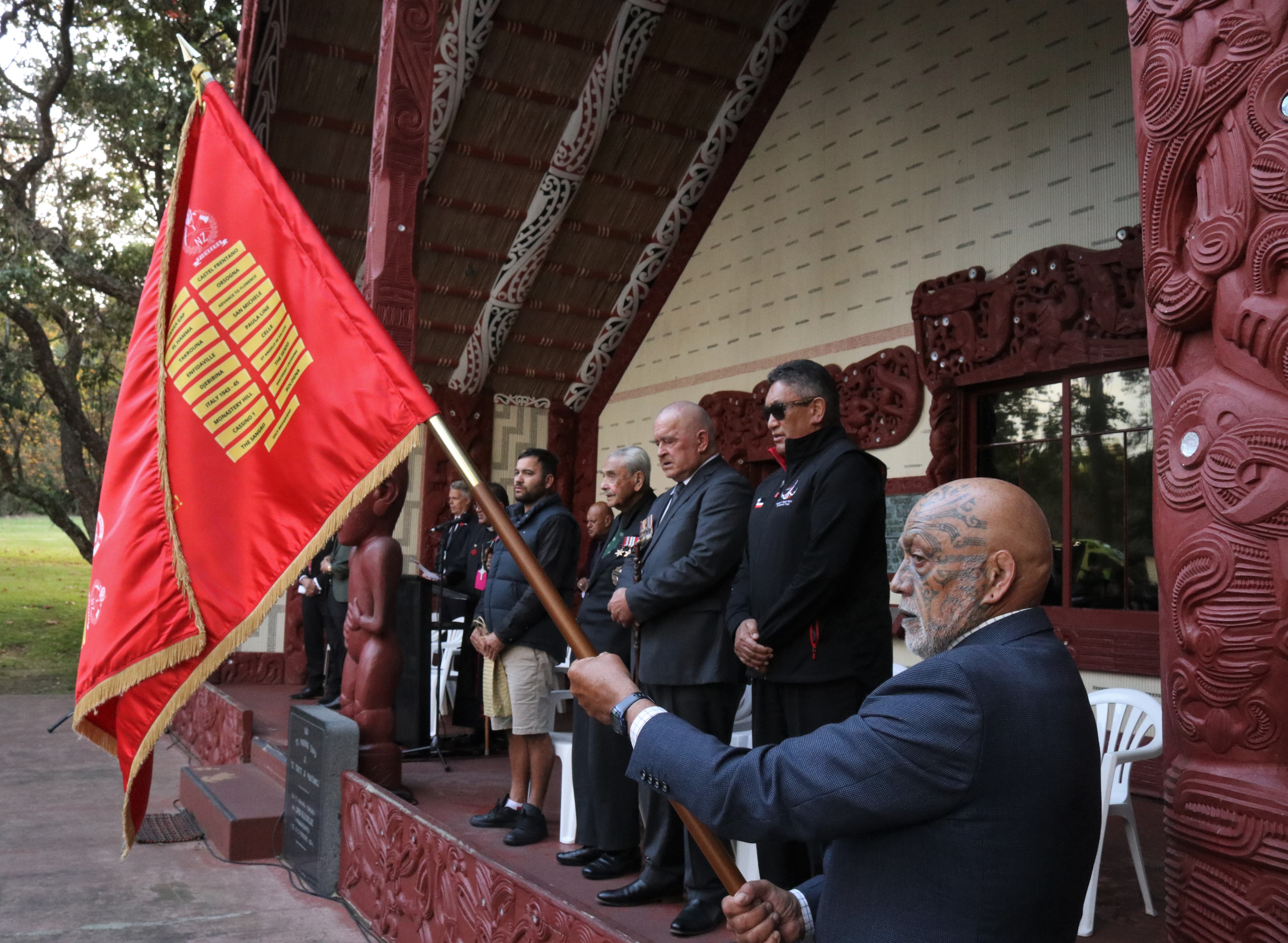 Tā (Sir) Robert “Bom” Gillies (fourth from right) at the Treaty Grounds’ Te Whare Rūnanga, flanked by grandson Te Whanoa Gillies, Bernard Henare, Hone Harawira and Harawira Pearless, holding the 28th Māori Battalion Battle Honours memorial flag. Photo / Peter de Graaf
