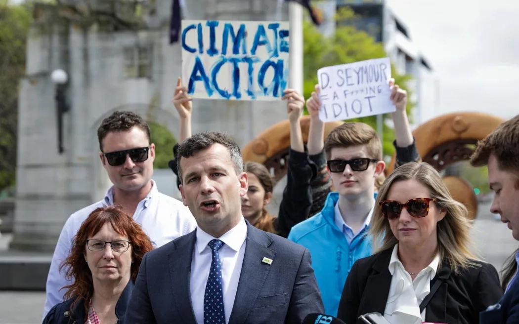 Teenagers protest in the background while Act leader David Seymour addresses media in Christchurch on Tuesday. Photo / Nathan Mckinnon, RNZ
