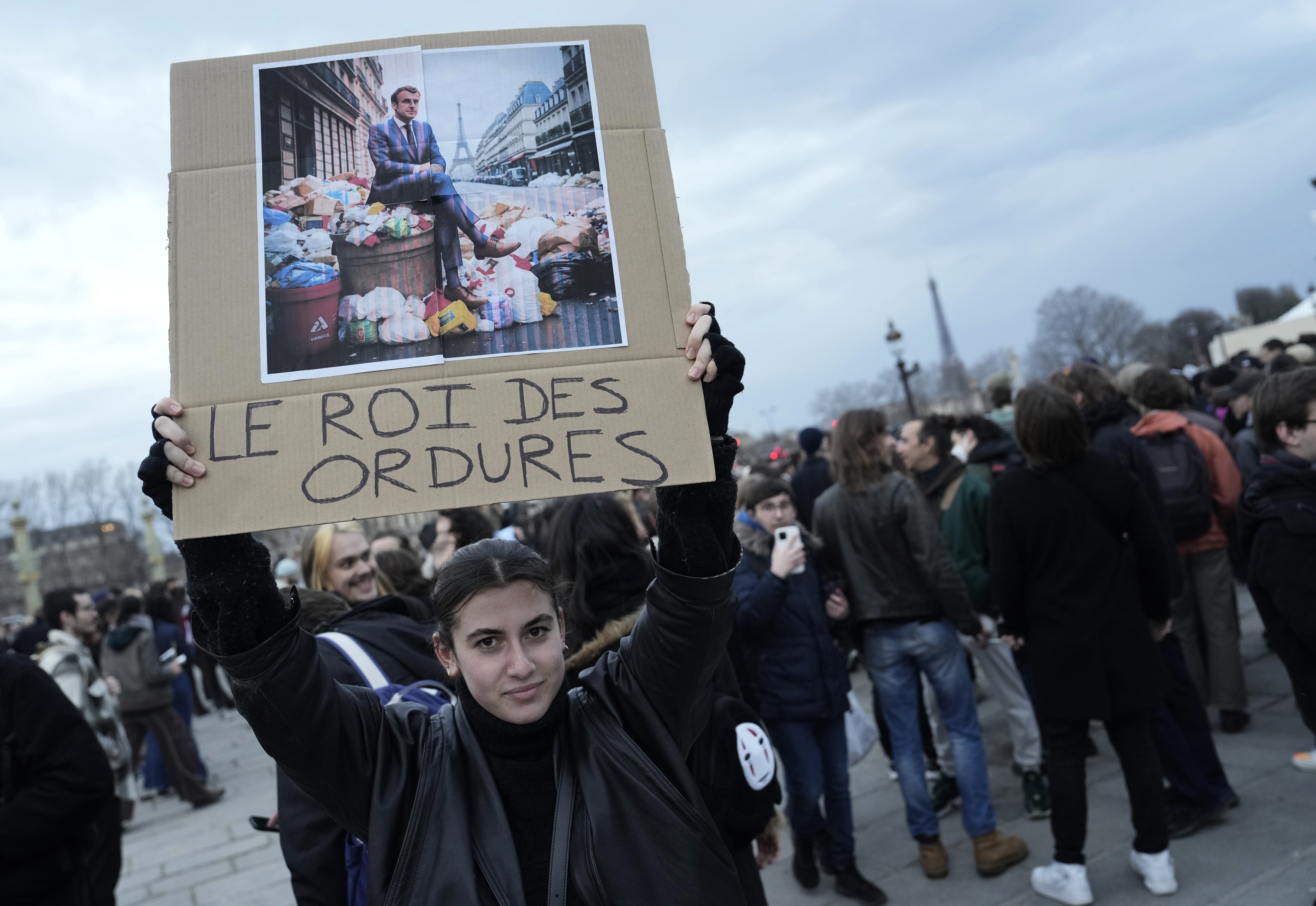 A woman holds a placard depicting French President Emmanuel Macron sitting on garbage cans that reads,