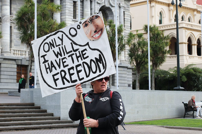A woman holds a banner showing New Zealand Prime Minister Jacinda Ardern with a swastika on her forehead and the letters CCP (Chinese Communist Party) on her cheek and the words 'Only I will have the freedom