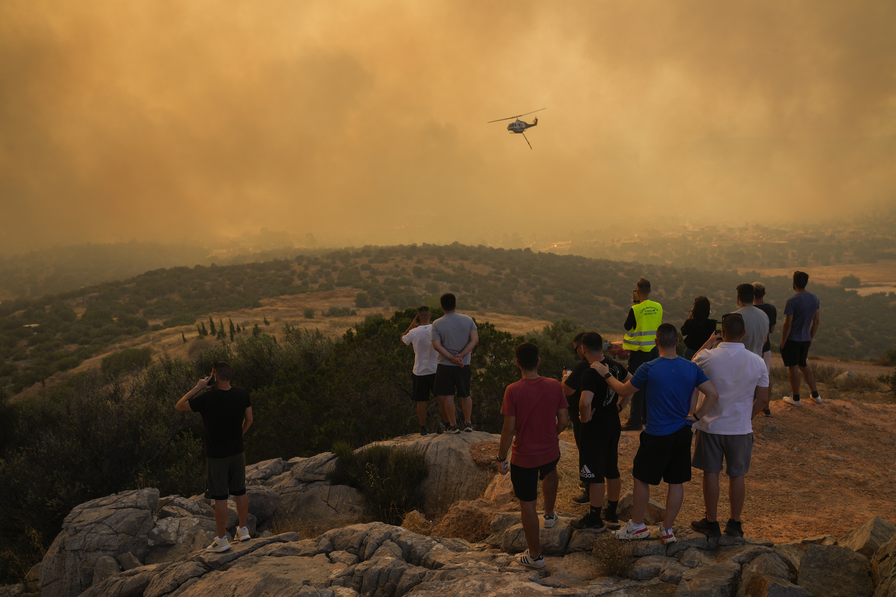 A fire-fighting helicopter flies through smoke as people look on in Mandra, west of Athens, on July 18. The world has likely just experienced its hottest July in recorded history. Photo / AP