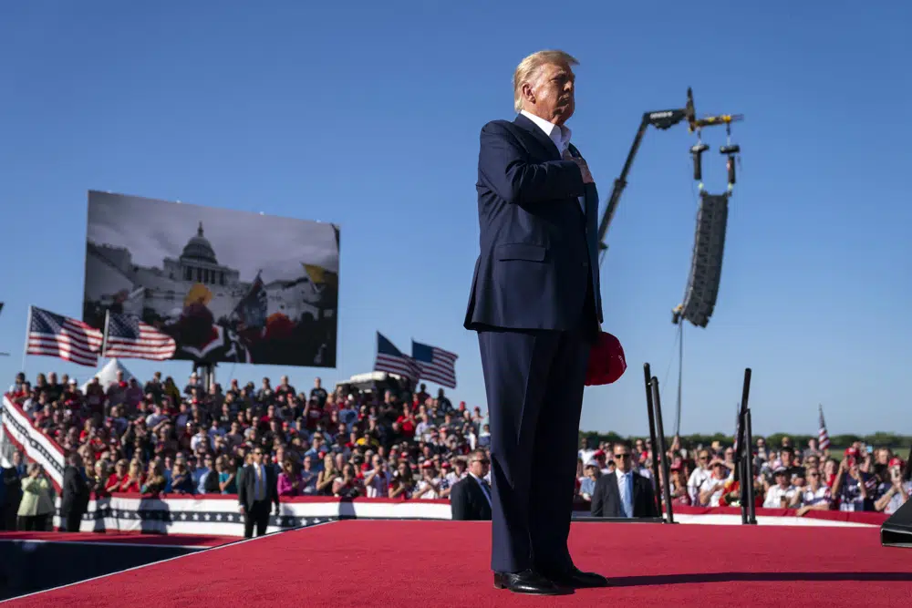 As footage from the January 6, 2021, insurrection at the US Capitol is displayed in the background, former president Donald Trump stands while a song, 
