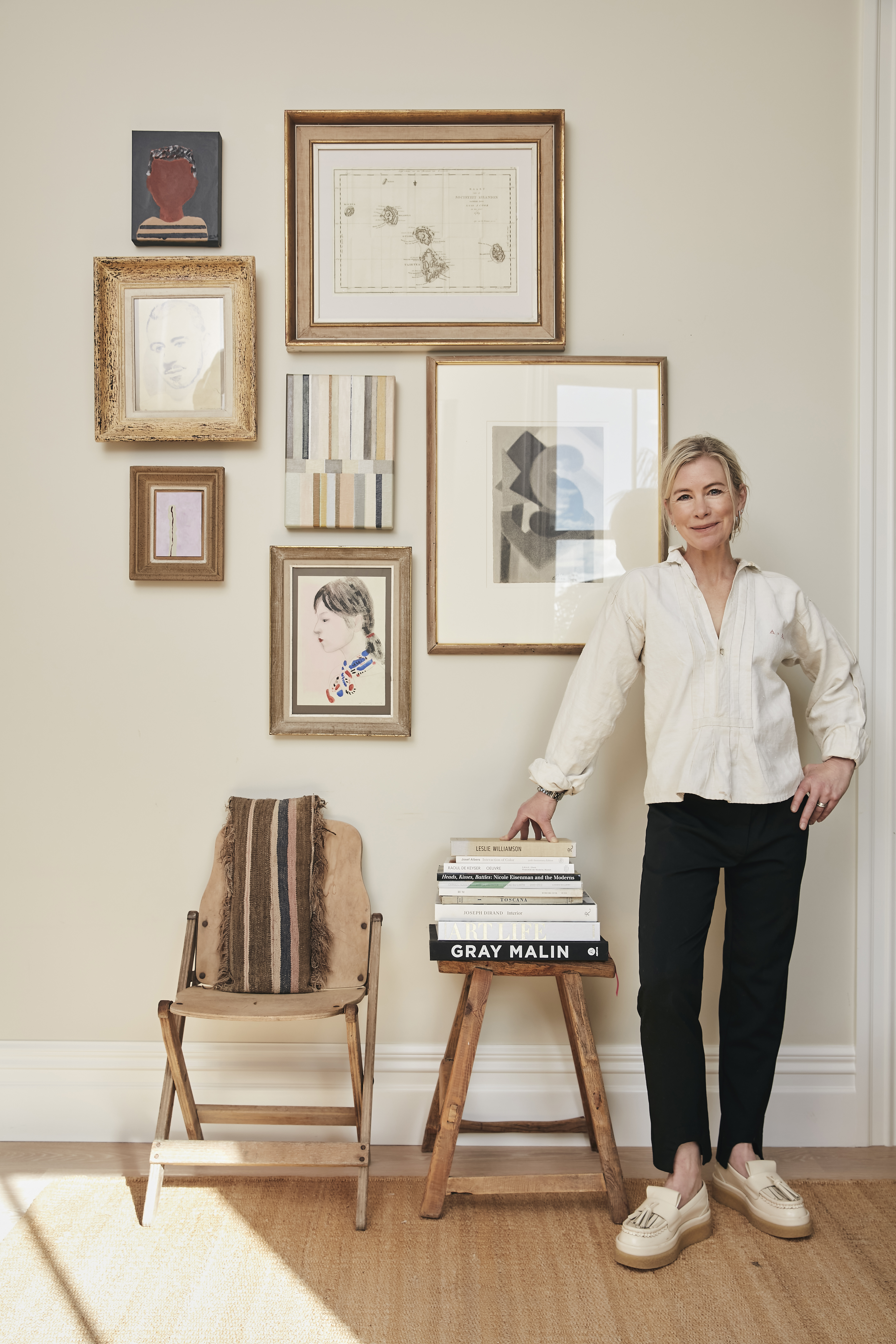 Founder of My Exhibition, Sara Bruce at home in front of her monochromatically toned artworks, including her own modernist paintings, other works were collected from local auctions and the portrait in a Montparnasse frame is from Paris, chair (far left) from auction, cushion from Babalogue and stool laden with art books from Flotsam & Jetsam. Photo / Babiche Martens