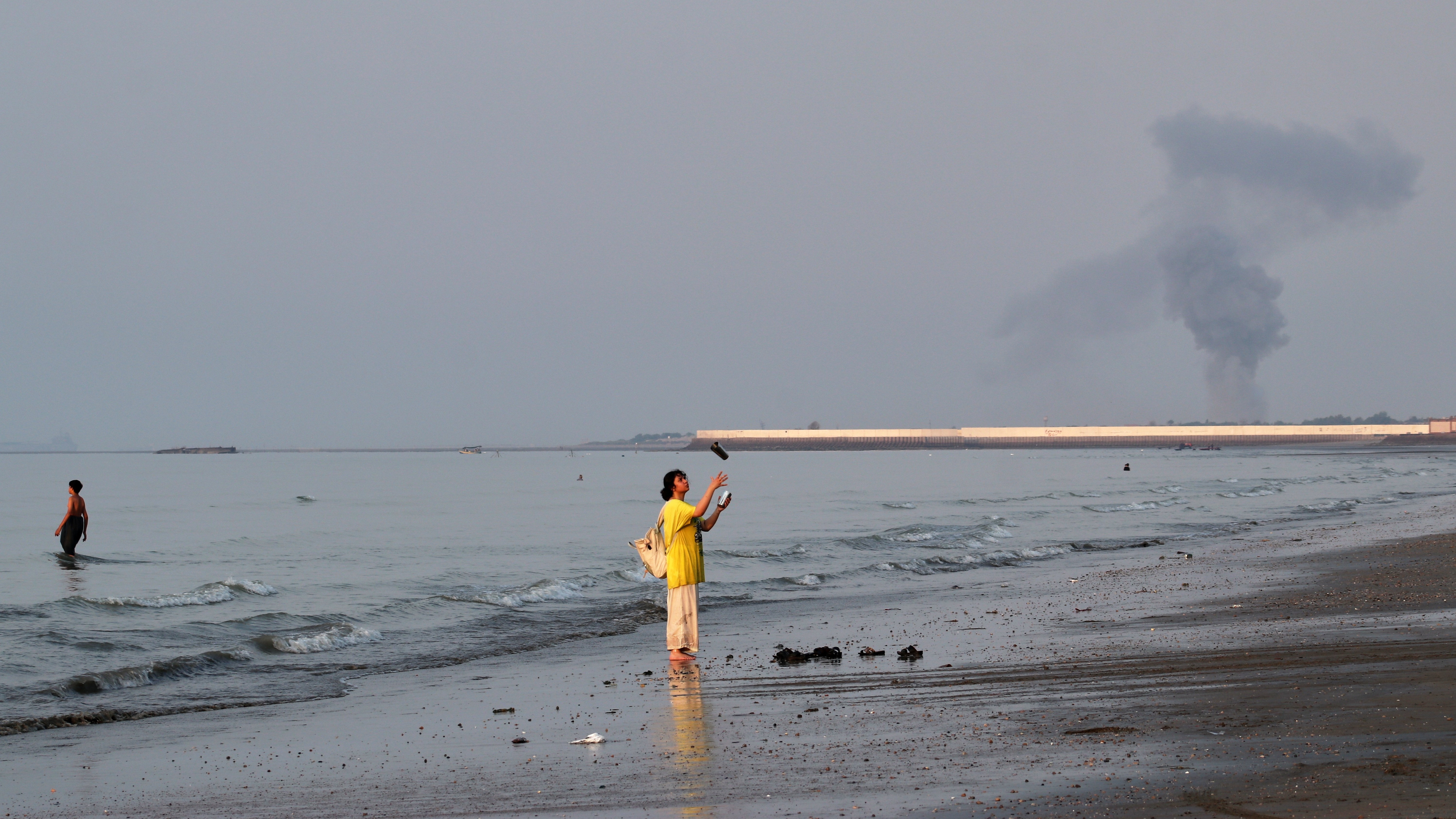A woman stands at the water's edge along the Strait of Hormuz as a plume of smoke rises in the background following an explosion, off Bandar Abbas, Iran.