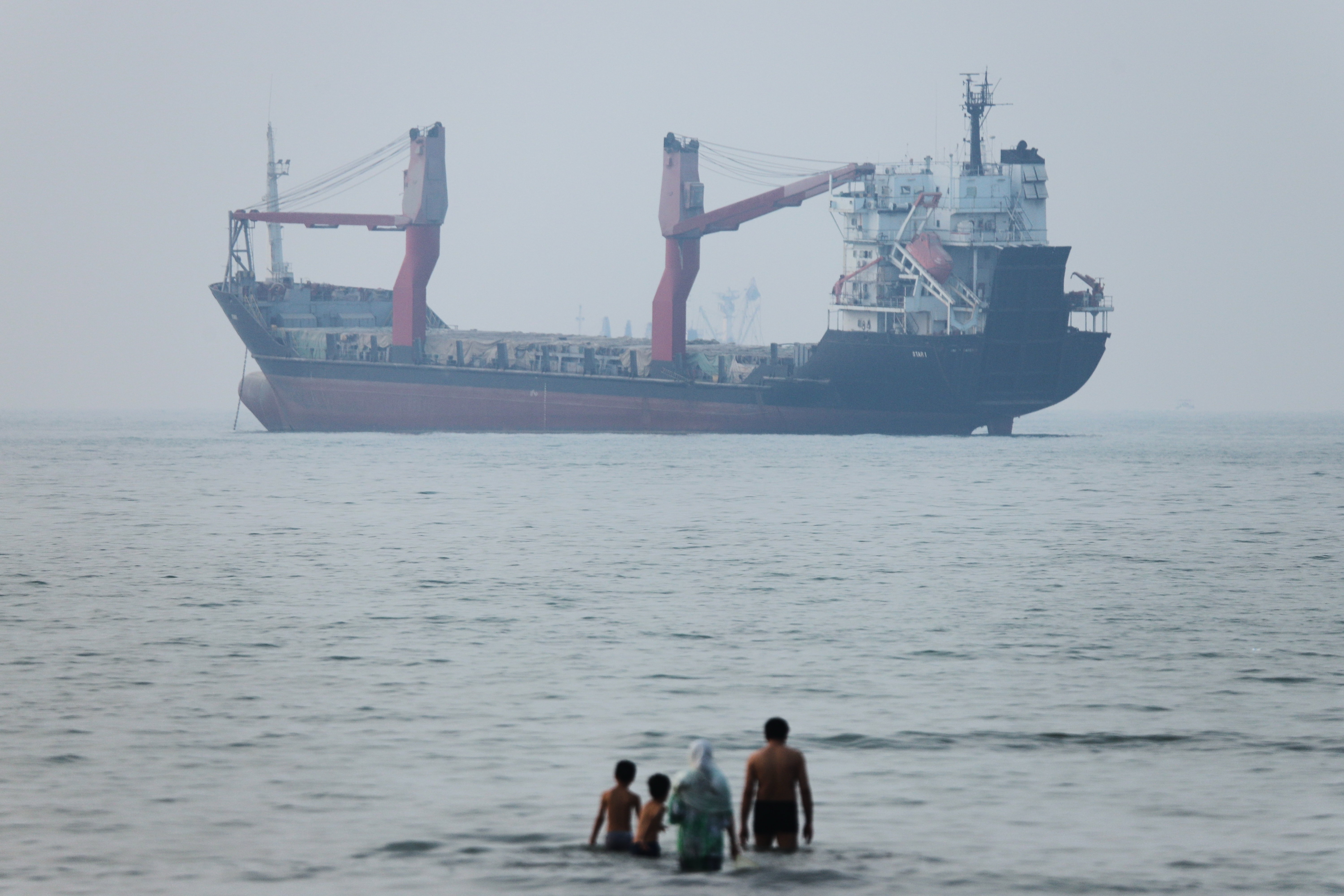 A group of people stands in shallow water as a cargo ship appears anchored in the Strait of Hormuz off Bandar Abbas, Iran, Tuesday, June 30, 2026.