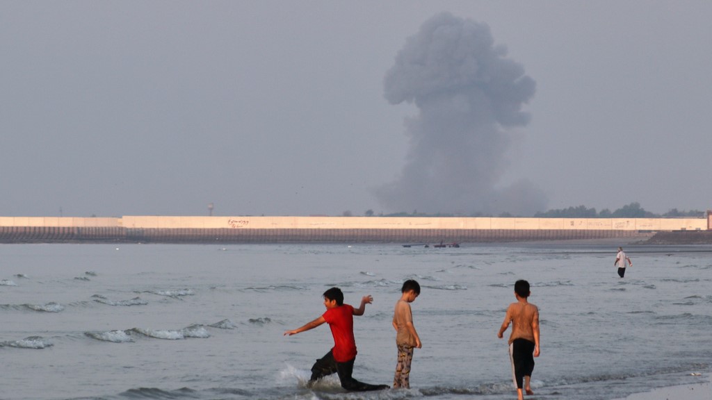 Three boys play in the shallow waters of the Strait of Hormuz, as a plume of smoke rises from an explosion in the background, off Bandar Abbas. (Source: Razieh Poudat/ISNA via AP)