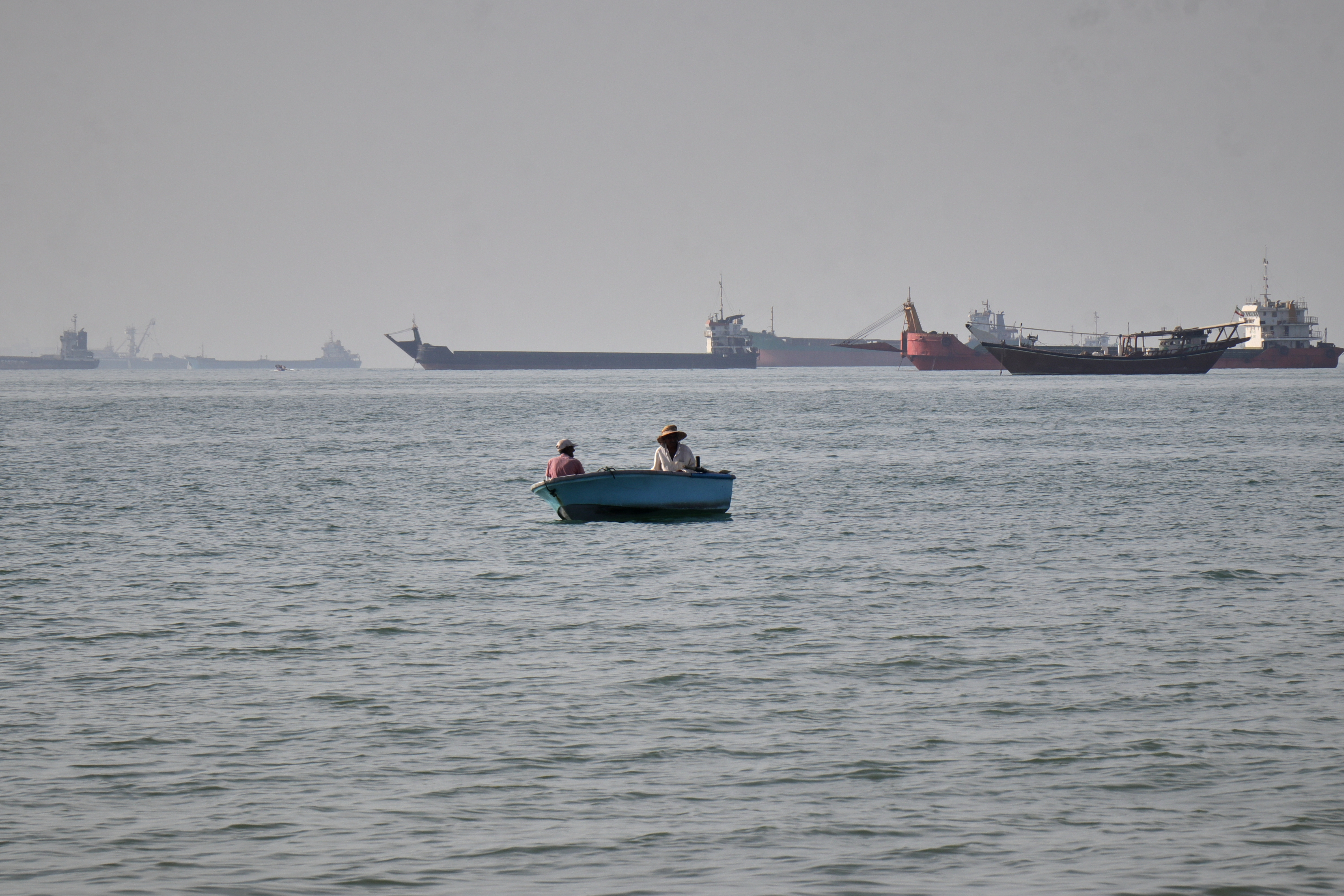 Two men sit in a small boat on the water as a mix of bulk carriers, cargo ships, and service vessels line the horizon in the Strait of Hormuz off Bandar Abbas, Iran, Monday, April 27, 2026.