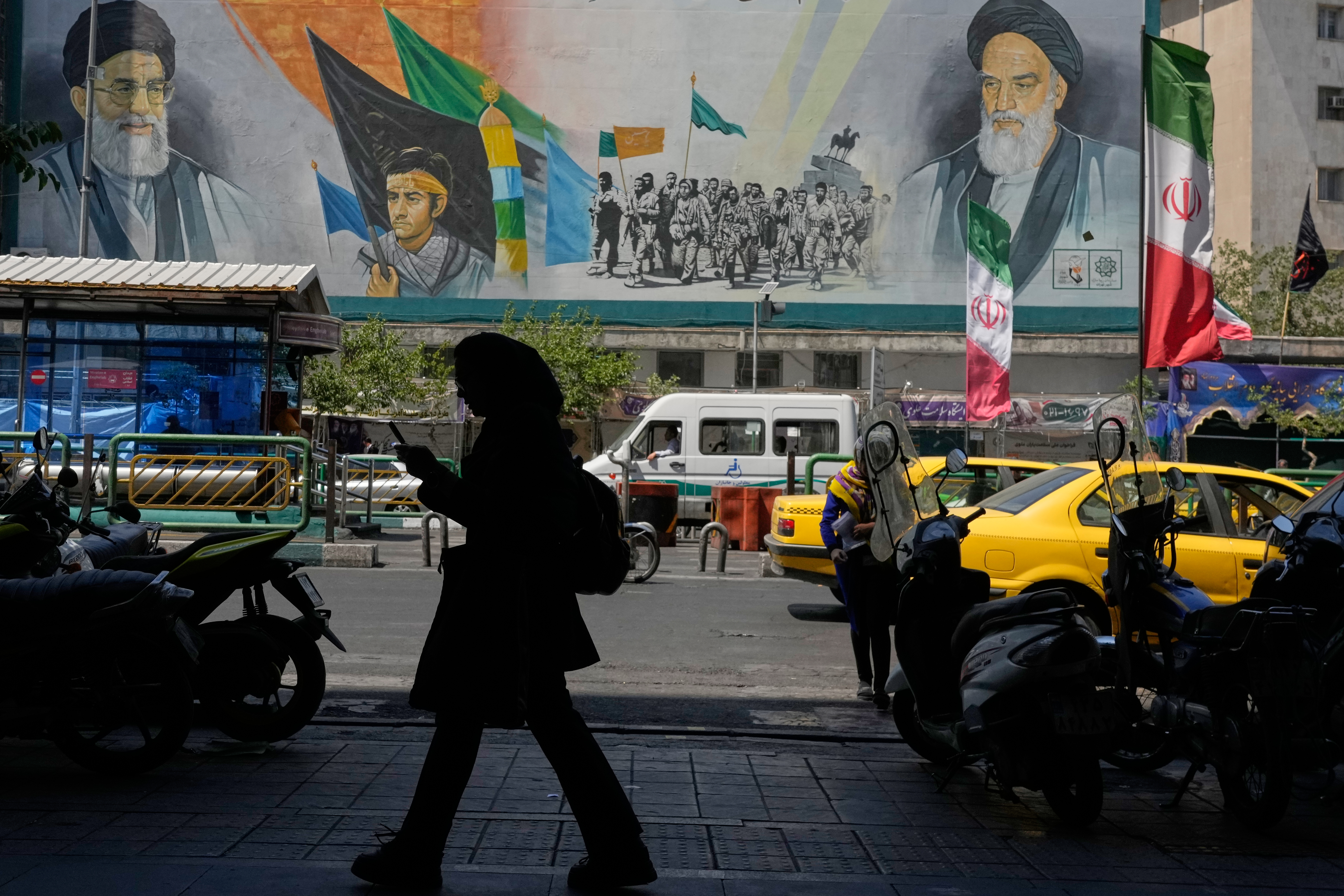 A woman walks on a sidewalk in front of a mural showing portraits of the late Iranian revolutionary founder Ayatollah Khomeini, right, the slain Supreme Leader Ayatollah Ali Khamenei