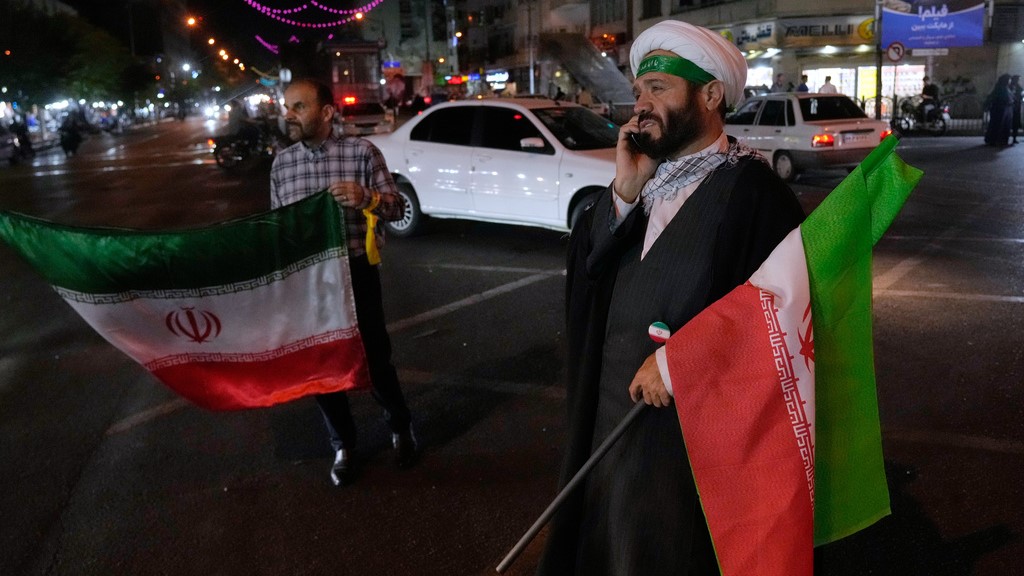 A cleric speaks on his cellphone holding an Iranian flags for a pro-government campaign in downtown Tehran, Iran, Friday, May 8, 2026.