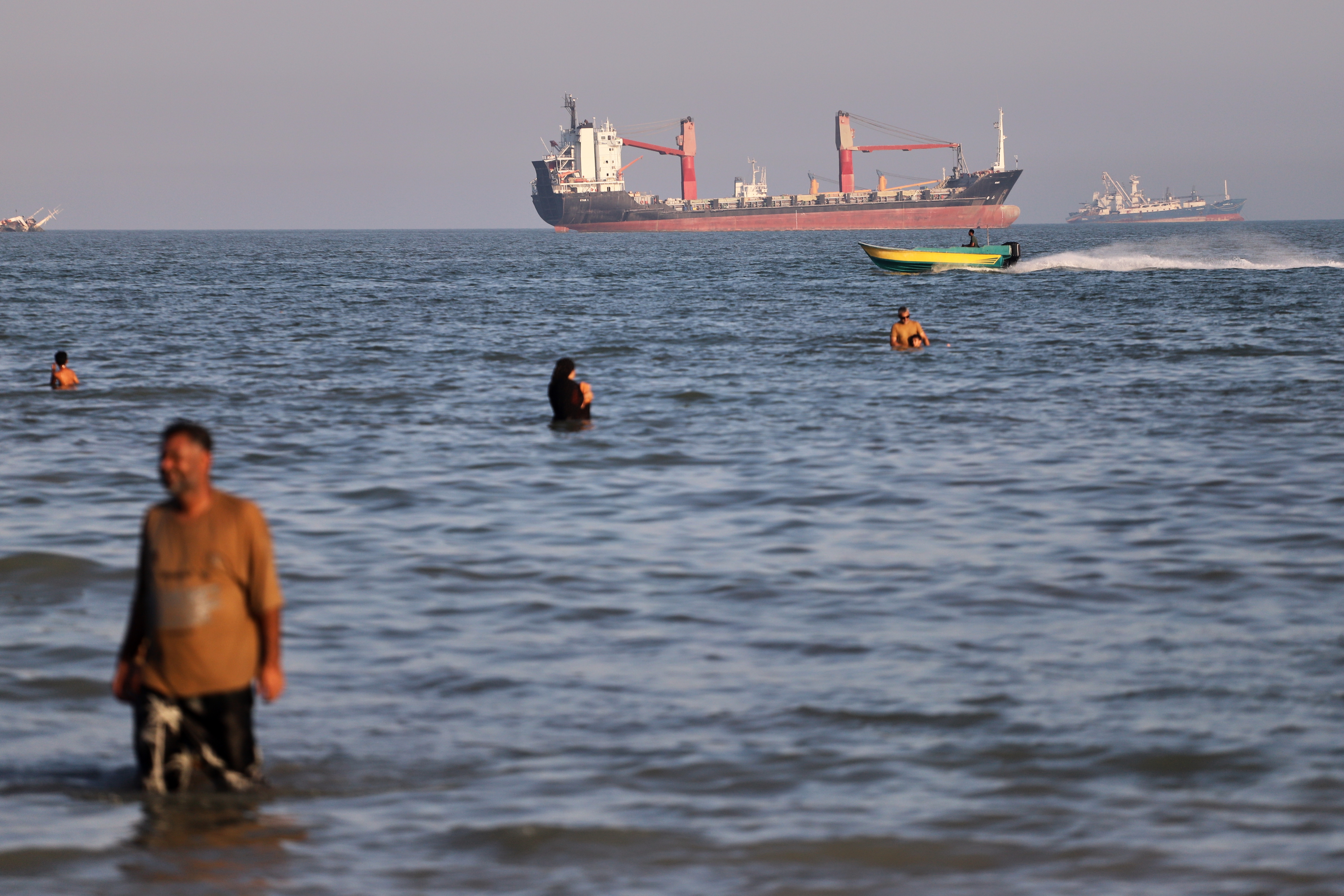 Residents swim in the waters of the Strait of Hormuz as a small motorboat passes cargo ships and other commercial vessels offshore near Bandar Abbas, Iran, Wednesday, June 17, 2026.