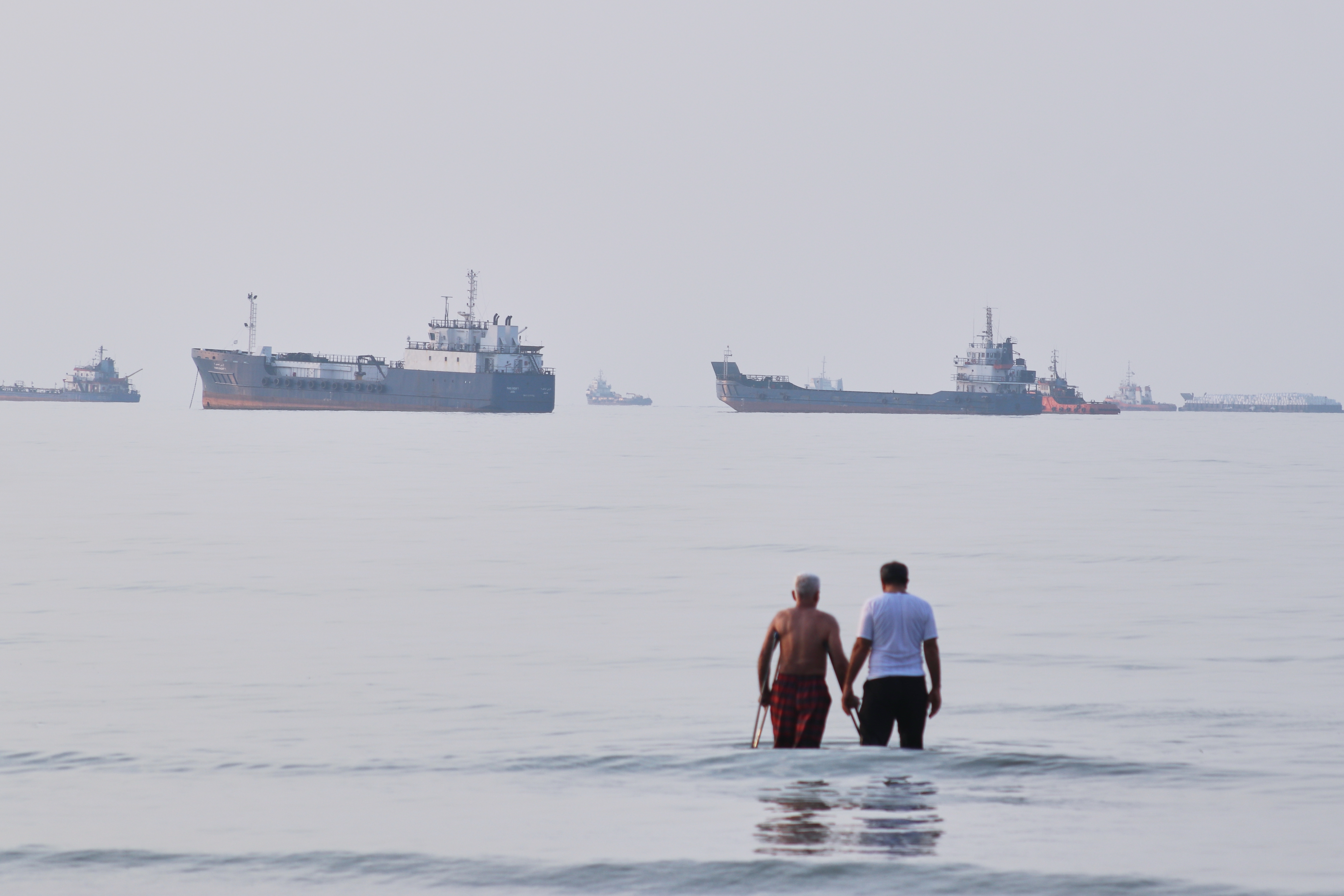 Two men wade in the waters of the Strait of Hormuz with vessels anchored in the background, off Bandar Abbas, Iran, Sunday, July 12, 2026.